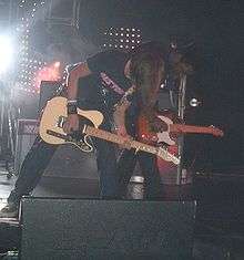 Two guitarists perform energetically side-by-side on a stage lit by white lights.