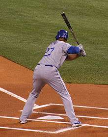 A man stands in a batter's box hunched over home plate. He is wearing a gray baseball uniform that reads "Kemp" and "27" on the back in blue block lettering and a blue batting helmet.