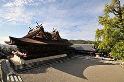 A large wooden building with two small gabled roofs put on top of the main roof and across the main ridge. There are forked roof finials on the top.
