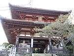 A two-storied wooden gate with white walls and faded red colored beams. There are two guardian statues in the side bays of the lower floor.