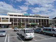 In front of a low and ivory building, a parking lot and cars are shown.