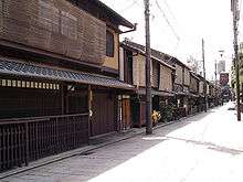 Wooden two-storied houses lining a small street. The upper stories' windows are covered.