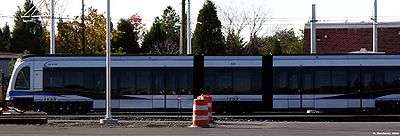 A blue and gray with black glass train stopped with overhead wires and trees visible.