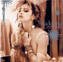 Madonna wearing a wedding dress and heart-shaped earrings is using one of her gloved hands to pull down on her pearl necklace.