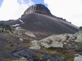 Rocky mountain with smooth steep flanks and a flat summit rising over rocks and sparse vegetation.