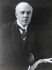 A balding, white-haired man wearing 1920s formal business attire (jacket and waistcoat with wing collar and tie) leans to his left on a table and looks directly at the viewer. His right hand rests in front of his left on the edge of the table next to some papers and a pen.