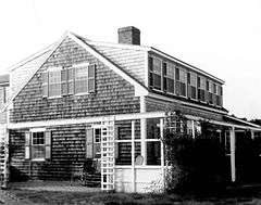 A modest Cape-style house with large flat-roof dormers on the front and back. There is a patio area on the side of the house, with an arbor-like structure separating it from the yard.