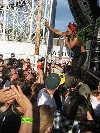 A dark-skinned woman wearing a black hat and black clothing holding a microphone while performing on stage to a crowd of people.  The performance is taking place outdoors and part of a rollercoaster is visible in the background.