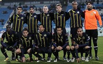 A modern association football team lines up for a pre-match photograph. Five players squat in front of a line of six, which stands. All of the players wear a dark blue uniform with yellow markings, apart from the player to the extreme right of the standing row, who wears an orange shirt with black shorts and socks. He also wears a pair of white gloves, indicating that he is the team's goalkeeper.