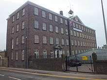 A four storey brick building, on the long side to the right there are 17 windows on each floor, on the short side there are only 3 window each floor. Over the central five windows is a triangular pediment surmounted by a cupola with a bell. Over the ground floor door, four bays from the near corner is the date 1790.