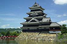 A large five-story castle tower with black wooden walls located on a platform of unhewn stones surrounded on two sides by water. The tower is connected to the lower structure.