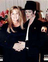 A man stands to the left of a woman who is smiling. He wears a black military jacket, under which is a white T-shirt. Atop his head is a black fedora. His left hand holds the right of the woman's. Both hands are pressed against the stomach of the woman, who is dressed in black.