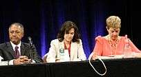 From left to right, an older man in a dark suit, a middle aged woman in a white blazer, and a middle aged woman in a salmon blouse sit at a long dark table in front of dark blue curtains.