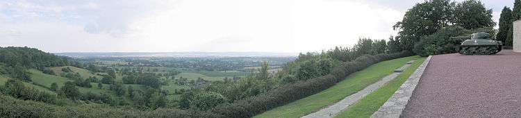 In the foreground tree-covered hills rising to the left and right frame a view over a valley. A tank and the edge of a building are on the rightmost hill. A flat plain of fields, trees and hedgerows fills the background, with a small hamlet visible in the middle-distance.