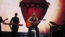 Image of a blonde woman sitting on a stool. She's wearing a black sleeveless shirt and black pants. She is holding an orange acoustic guitar in her hands. A microphone stands before her. Other instruments and musicians can be seen in the background.