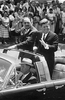 (L-R) English: Motorcade for President Mohammad Ayub Khan of Pakistan. In open car (Lincoln-Mercury Continental with bubble top): Secret Service agent William Greer (driving); Military Aide to the President General Chester V. Clifton (front seat, center); Secret Service Agent Gerald "Jerry" Behn (front seat, right; partially hidden); President Mohammad Ayub Khan (standing); President John F. Kennedy (standing). Crowd watching. 14th Street, Washington, D.C.