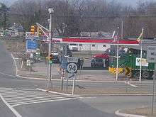 A road approaching a signalized intersection with a sign at a short ramp reading north Route 94 with an arrow pointing to the upper right. In the distance, a gas station can be seen along with road signs for north Route 15 and south Route 94.