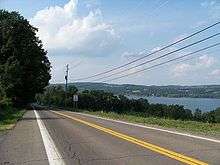 A two-lane highway runs alongside a large lake. The road and the lake are separated by a row of trees and small brush.