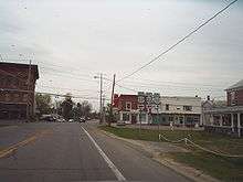 A two-lane highway approaches an intersection with another highway regulated by a blinking signal. Small, mostly two-story buildings surround the rural junction. A sign assembly indicates that NY 22 northbound and US 11 southbound turn left while US 11 northbound is straight ahead.