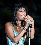 A woman wearing earrings, a bracelet and a multi-colored spaghetti strapped dress, with both hands folded over a microphone.