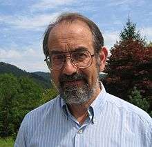 A man in late middle-age with a greying black beard and thinning hair, wearing an open-necked casual blue shirt and large thin-rimmed glasses