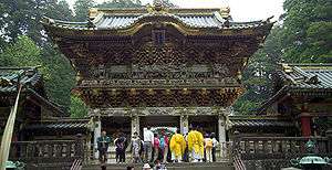 A two-storied, decorated wooden gate where the upper story extends over the lower. There is a bulging gable at the center under which a board with characters is attached.