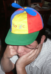 Asian man in his twenties wearing a blue, green, yellow and red propeller hat that says "Noogle"