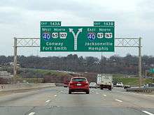 Traffic flows down a four-lane Interstate highway underneath a large sign indicating traffic exits