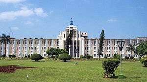 A wide four storied building with landscaped lawn and garden in the foreground