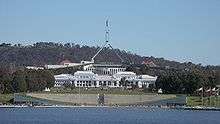 Bright white classical building in front of larger, modernistic beige, gray, and green building.  Two metal spires stretch from the top of the larger building in a trangular shape; near the top, before the two connect, they bend straight up, supporting a flagpole and flag.