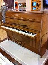 An image of a medium-sized brown upright piano in a glass case. The piano keys are exposed.