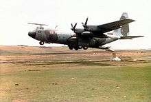A Royal Air Force C-130 Hercules  military plane. Its propellers attach to its wings, and it appears to be hovering just above a barren field. From the back of the aircraft, a package is being dropped onto the field from an open rear gangway.