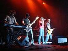 Four people on a stage. The first one with a plaid shirt and blue jeans, playing a keyboard. The second one wearing a black cap, a striped shirt and blue jeans, playing a bass guitar. The third one is a female wearing a gray hat, a pink shirt and blue jeans, holding a microphone. The fourth one, is wearing a gray shirt and blue jeans and is playing an electric guitar.