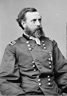 Head and torso of a white man with a full beard, wearing a partially unbuttoned double-breasted military jacket, a handkerchief tied around his neck, and a single medal pinned to his left breast.