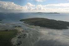 Two dark green areas of land are separated from one another by a shallow and narrow strait. There are small islands in the distance under white clouds.