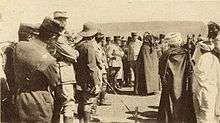 French and Moroccan men crowd around the central figures of Hassan and Poeymirau, standing in discussion