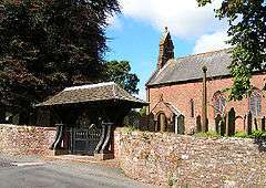 The western part of a church seen from the southwest with a west bellcote and a large roofed lychgate in the foreground