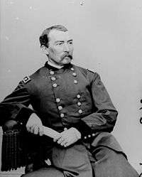 Man with moustache sitting down with arm on table in uniform with two columns of buttons