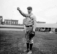 A black-and-white photograph of a man wearing an old-style baseball uniform and a newsie cap holding a baseball in his outstretched hand