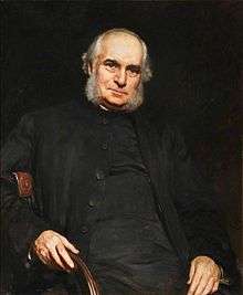A balding man with grey/white sideburns, wearing a dark jacket over a dark robe, sitting in a chair