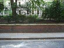 A brick wall topped by metal railings stands at the side of a tarmac road. Behind the wall, grass and trees are visible, growing at the height of the top of the wall.