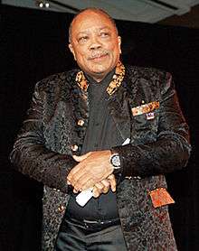 Quincy Jones, a plump bald African American man with a grey moustache and wry smile. He is elegantly dressed in a black brocade jacket with patterned collar over a black shirt.
