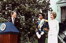 President Reagan wearing a suit and tie stands at a podium and turns to smile at Mrs Reagan, who is wearing a white outfit, and Jackson, who is wearing a white shirt with a blue jacket and a yellow strap across his chest.