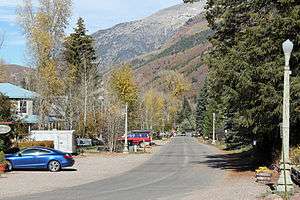 A street lined with streetlights and houses on large lots sheltered by trees, leading towards tall mountain slopes in the distance