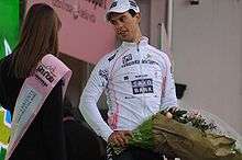 A man in his early twenties wearing a white cycling jersey and cap holding a bouquet of flowers looks to his right at a woman, whose face is not visible.