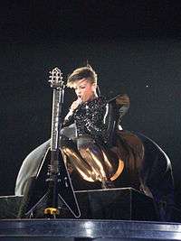 A woman wearing a black outfit. She is sitting on a chair and holding a guitar