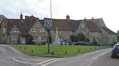 Street scene. Triangular area of grass with village sign on wooden post and stone cross behind. Stone houses with tiled roofs n the background.