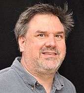 A brown haired man in a gray shirt looks into the camera while standing against black background.