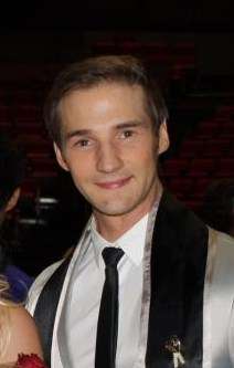 A photograph of a blond man wearing a silver suit, a white dress shirt, and a black necktie while looking at the viewer and smiling