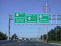 A six-lane divided highway approaching an intersection with a set of three green signs over the roadway. The left sign reads Route 70 west Lakehurst Camden keep left, the middle sign reads north Route 34 to Garden State Parkway Matawan, and the right sign reads Route 35 north Belmar next right.